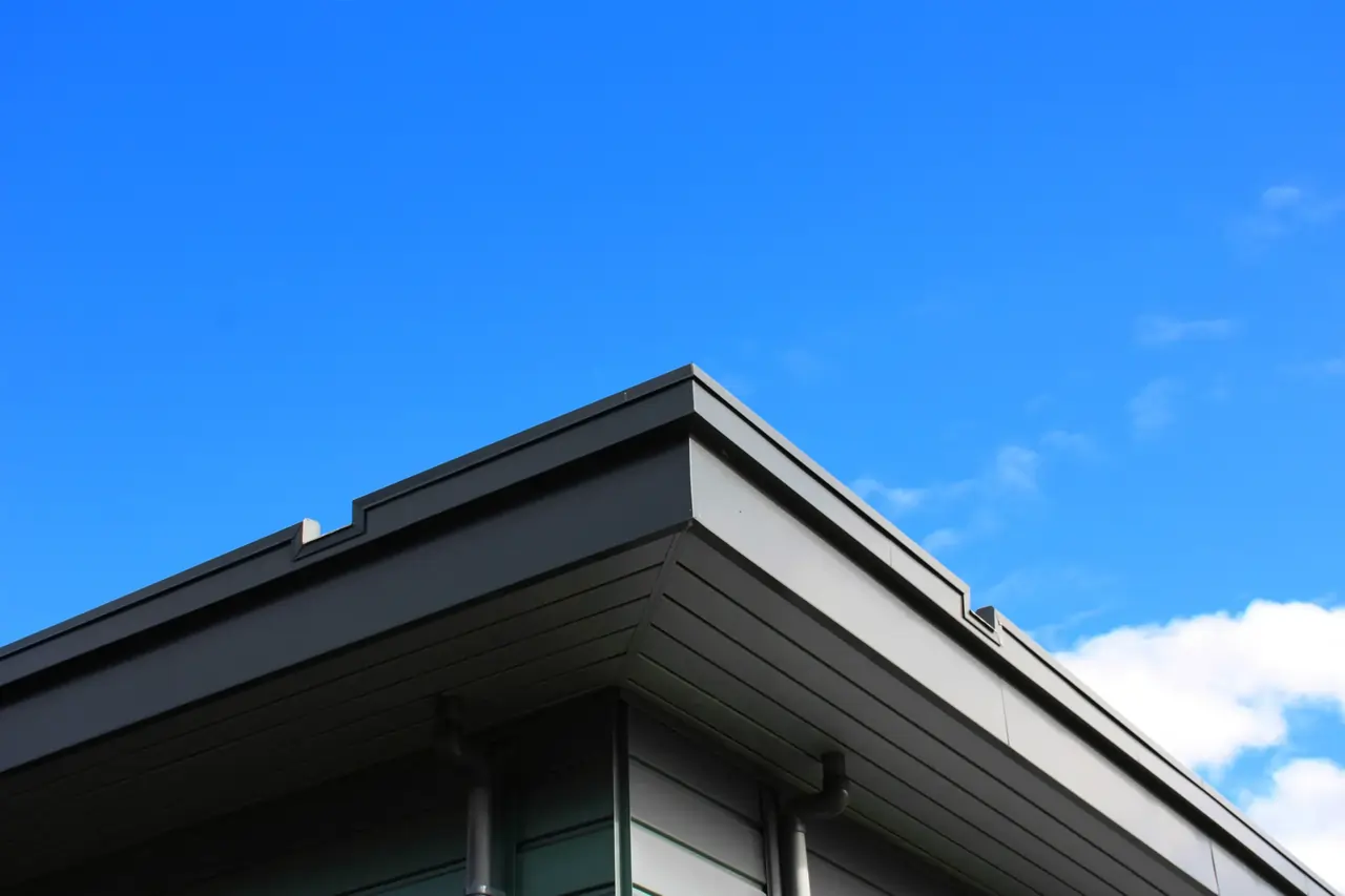 Modern gebouw met een strak afgewerkt plat dak en grijze gevelpanelen, met zicht op een aangrenzend hellend dak.