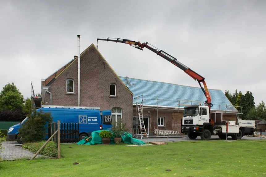 Dakwerken De Haes voert dakwerken uit op een woning in Sint-Katelijne-Waver, met een kraanwagen die materialen op het dak plaatst. Op de achtergrond is een blauwe bestelwagen van het bedrijf zichtbaar