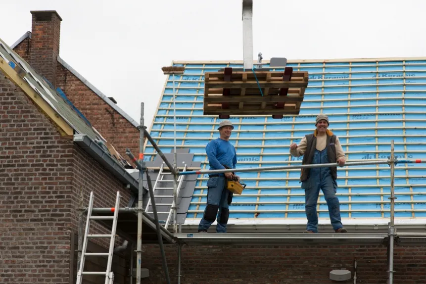 Twee dakwerkers van Dakwerken De Haes aan het werk op een dak in Sint-Katelijne-Waver, met dakisolatie en houten latten. Een pallet met materialen wordt via een kraan omhooggehesen.